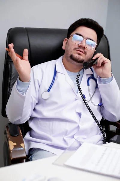 Ένας Νεαρός Γιατρός Κάθεται Στην Κλινική Και Μιλάει Στο Τηλέφωνο — Φωτογραφία Αρχείου
