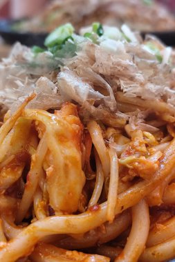 Karıştırılmış Japon udon eriştesi ve sebzeler, üzerine bonito gevreği ve soğan konmuş, cızırdayan bir demir tabakta servis ediliyor. Yemek çubuklarıyla sıcak bir yemek ortamında yakalandılar..