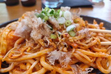 Karıştırılmış Japon udon eriştesi ve sebzeler, üzerine bonito gevreği ve soğan konmuş, cızırdayan bir demir tabakta servis ediliyor. Yemek çubuklarıyla sıcak bir yemek ortamında yakalandılar..