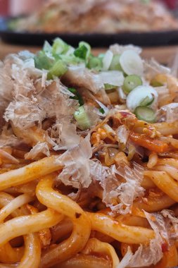 Karıştırılmış Japon udon eriştesi ve sebzeler, üzerine bonito gevreği ve soğan konmuş, cızırdayan bir demir tabakta servis ediliyor. Yemek çubuklarıyla sıcak bir yemek ortamında yakalandılar..