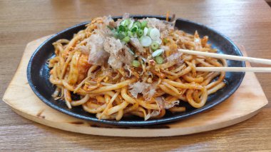 Karıştırılmış Japon udon eriştesi ve sebzeler, üzerine bonito gevreği ve soğan konmuş, cızırdayan bir demir tabakta servis ediliyor. Yemek çubuklarıyla sıcak bir yemek ortamında yakalandılar..