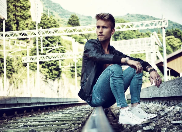 Attractive young man sitting on railroad, wearing black leather jacket, sneakers and jeans, looking away, at daytime