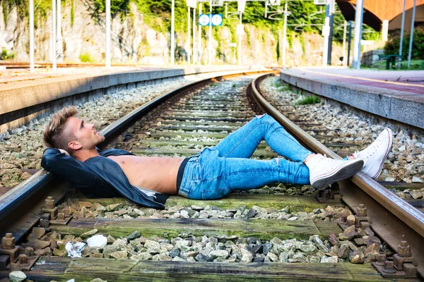 Attractive young man laying on railroad, wearing black leather jacket, sneakers and jeans, at daytime