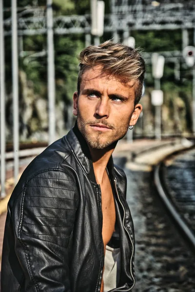 Attractive young man sitting on railroad, wearing black leather jacket looking away, at daytime