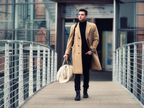 Confident young man walking in fashionable trench coat through city street. Holding a bag