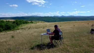 Dizüstü bilgisayarlı aktif ve mobil grafik tasarımcısı katlanabilir bir sandalyede oturuyor ve açık havada dizüstü bilgisayar ve grafik tabletle çalışıyor.