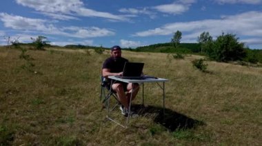 Dizüstü bilgisayarlı aktif ve mobil grafik tasarımcısı katlanabilir bir sandalyede oturuyor ve açık havada dizüstü bilgisayar ve grafik tabletle çalışıyor.