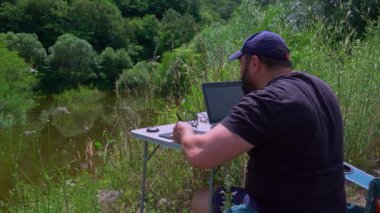 Dizüstü bilgisayarlı aktif ve mobil grafik tasarımcısı katlanabilir bir sandalyede oturuyor ve açık havada dizüstü bilgisayar ve grafik tabletle çalışıyor.