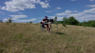 Dizüstü bilgisayarı olan aktif ve gezgin bir adam sandalyede oturuyor, kırsal kesimde ve ağ kuruyor.