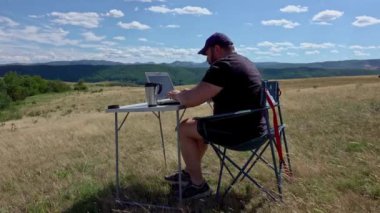 Dizüstü bilgisayarı olan aktif ve gezgin bir adam sandalyede oturuyor, kırsal kesimde ve ağ kuruyor.
