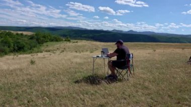 Dizüstü bilgisayarı olan aktif ve gezgin bir adam sandalyede oturuyor, kırsal kesimde ve ağ kuruyor.