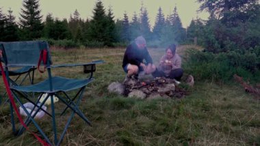 Erkek ve kadın, yürüyüşçüler şafakta ormanda kamp ateşinde yemek hazırlıyorlar. Yaz gecesi.
