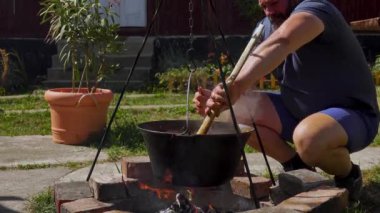 Geleneksel Romen zakuskasının ahşap bir ateşin üzerinde bir kazanda hazırlandığını gösteren kırsal bir sahne. Bu açık hava pişirme süreci doğal malzemelerin ve geleneksel yöntemlerin kullanımını vurguluyor ve bu da zengin ve lezzetli bir yayılımla sonuçlanıyor..