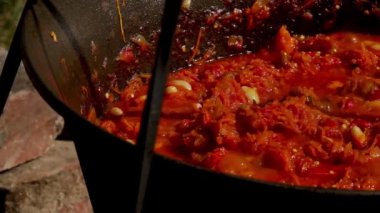 Geleneksel Romen zakuskasının ahşap bir ateşin üzerinde bir kazanda hazırlandığını gösteren kırsal bir sahne. Bu açık hava pişirme süreci doğal malzemelerin ve geleneksel yöntemlerin kullanımını vurguluyor ve bu da zengin ve lezzetli bir yayılımla sonuçlanıyor..