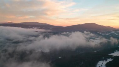 Drone flight among beautiful low clouds and mists creeping over the snow-covered mountain slopes, the Rhodopes in Bulgaria at winter sunrise.