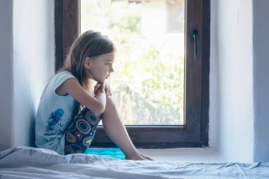 Vintage portrait of a beautiful little girl with bare feet and pensive gaze sitting near a window inside a room	