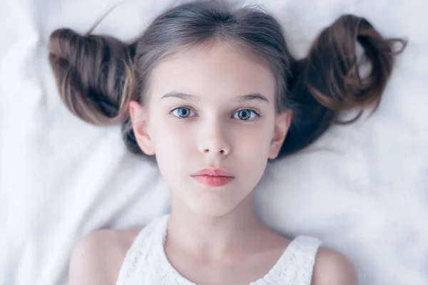 Close up shot of beautiful blonde caucasian little girl with fashionable hairstyle, lying on a bed in a white dress, looking calm and dreamy	