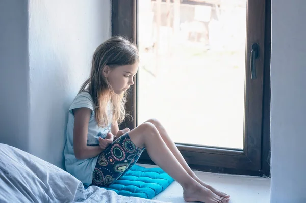 Vintage portrait of a beautiful little girl with bare feet and pensive gaze sitting near a window inside a room	