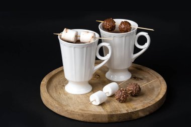 Two beautiful white porcelain cups of hot Chocolate drink with marshmallows and chocolates skewers over wooden table on black background. Winter time. Holiday concept.	