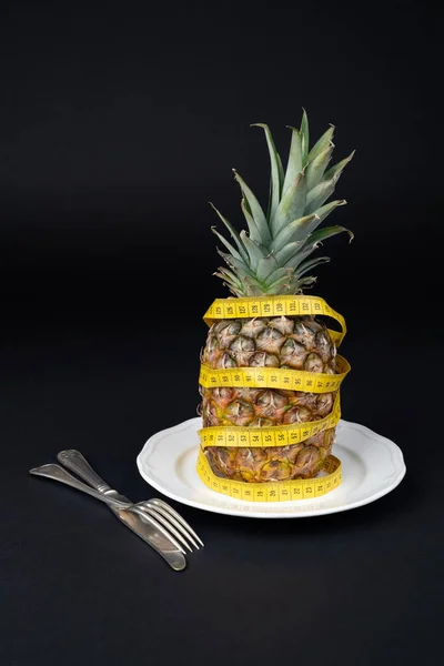 Ripe pineapple in a white plate wrapped with a tape measure on a dark background. Fitness concept, diet, healthy lifestyle.