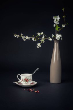 Güzel bir porselen çay fincanı, çay aksesuarları ve siyah arkaplanda çiçek açan bir dal ile dolu bir hayat.