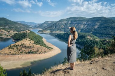 Rear view of a young long-haired woman enjoying the view of one of most picturesque meander of Arda river surounded by mountain slopes of Rhodopi Mountains, Bulgaria	 clipart