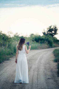 Beyaz elbiseli, uzun saçlı bir kadının gün batımından önce elinde lavanta buketi buketi ile yolda yürüdüğünü hayal edin.