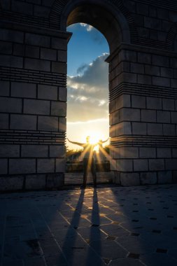 Silhouette of a woman sending the last rays of the sun at sunset, standing under the arch of an ancient building, part of the ruins of the Great Basilica near Pliska, Bulgaria.	