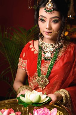 Çok güzel bir Hintli gelinin portresi. Lüks bir gelin kostümü içinde. Makyajlı ve stüdyoda ışık saçan bir nilüfer tutan ağır mücevherler. Yaşam tarzı Düğün modası.