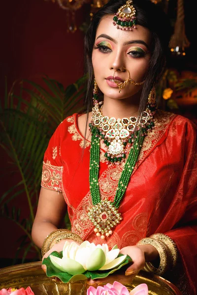Portrait of very beautiful young Indian bride in luxurious bridal costume with makeup and heavy jewellery holding a lotus in studio lighting indoor. Lifestyle Wedding fashion.