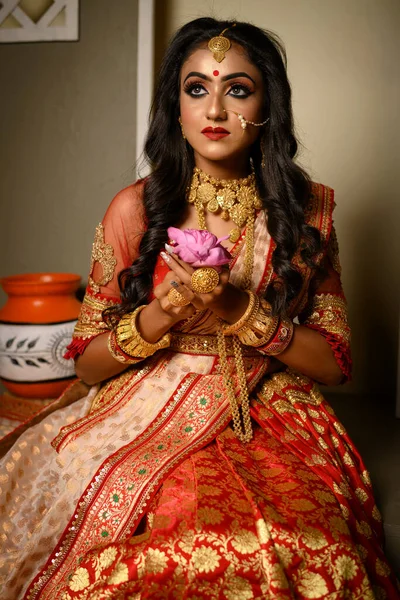 Portrait of pretty young Indian girl wearing traditional saree, gold jewellery and bangles holding flowers in her hands in studio lighting indoor. Indian culture, occasion, religion and fashion.