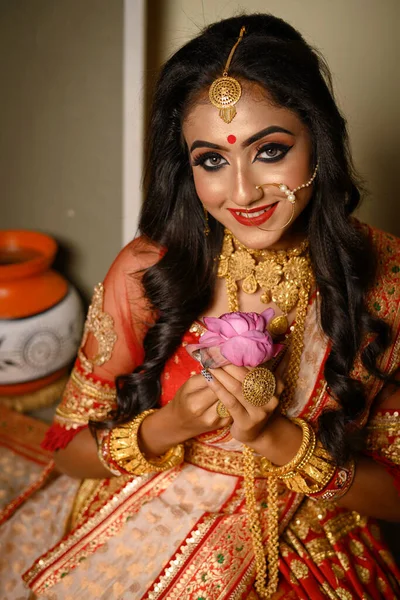 Portrait of pretty young Indian girl wearing traditional saree, gold jewellery and bangles holding flowers in her hands in studio lighting indoor. Indian culture, occasion, religion and fashion.