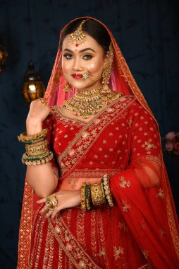 Çekici Hintli gelin, stüdyo ışıklarında ağır altın mücevherler ve peçeli geleneksel kırmızı gelinlik giyerek nazikçe gülümsüyor. Düğün modası ve yaşam tarzı.