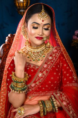 Çekici Hintli gelin, geleneksel kırmızı gelinlik içinde ağır altın mücevherler ve peçeyle stüdyo ışıklarında nazikçe gülümseyen bir sandalyede oturuyor. Düğün modası ve yaşam tarzı.