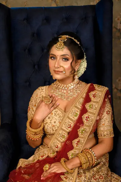 Stunning Indian bride dressed in traditional bridal lehenga with heavy gold jewellery and veil sitting in a chair with classic vintage interior in studio lighting. Wedding fashion.