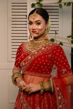 Geleneksel kırmızı lehenga ile süslenmiş göz kamaştırıcı Hintli gelin ağır altın takılar ve duvak takıyor ve stüdyo ışıklarında şefkatle gülümsüyor. Düğün modası ve yaşam tarzı.