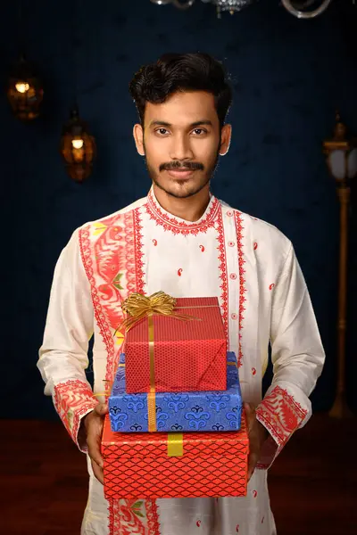 stock image Portrait of a young and handsome man, clad in a traditional kurta pyjama, holding gift boxes in hands under studio lighting. Indian fashion, events and lifestyle.