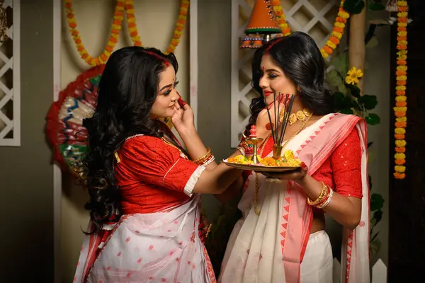 stock image Indian women, adorned in traditional sarees and gold jewellery, including bangles, holding a plate filled with religious offerings. Indian festival, culture, occasion, religion and ethnic fashion.