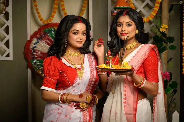 stock image Indian women, adorned in traditional sarees and gold jewellery, including bangles, holding a plate filled with religious offerings. Indian festival, culture, occasion, religion and ethnic fashion.