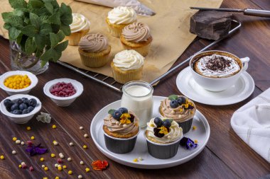 Taze pişmiş kek ve kahve. Kremalı ve çikolatalı kapuçino. Çeşit çeşit yenilebilir çiçekler ve küçük orman meyveleriyle süslenmiş kekler. Tahta zeminde sunum hazırlandı.