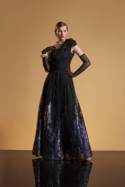 Saf eldivenleri, pırıl pırıl eteği ve inci aksesuarları olan, tek omuzlu, siyah gece elbisesi giyen kendine güvenen bir kadın. Stüdyoda bej arka planda poz veriyor. Ebedi zarafet ve modaya uygun bir bakış..