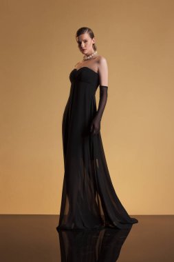 Sofistike bir kadın uzun siyah eldivenler ve inci kolyeyle süslenmiş straplez siyah bir gece elbisesi içinde bej renkli bir stüdyo arkaplanına poz veriyor. Görüntü, zamansız zarafeti, yüksek modayı yakalar. Siyah elbiseli güzel bir kadın.