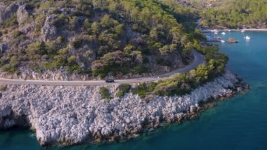 Tropikal Akdeniz iklimi, dağlar ve deniz iç içe geçmiş. İHA kamera siyah arabayı izleme sahnesi gibi takip ediyor. Turkuaz denizli yeşil çam ağaçlarının üzerinde uçuyor