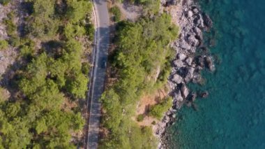 Deniz kenarındaki asfalt yol, çam ormanları ve yolda giden siyah araba. Drone arabayı takip ediyor. İHA 'yla üstten çekim. Akdeniz iklimi, dağlar ve deniz iç içe geçmiş. 
