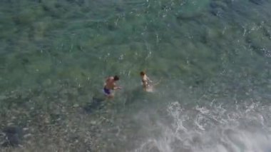 Neşeli genç bir çift, berrak mavi sularda su sıçratarak ve gülerek birlikte denizin tadını çıkarıyor. Hava görüntüsü romantizm, neşe ve yaz tatili titreşimlerini yakalıyor.