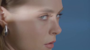Işıl ışıl cildi ve yoğun bakışları olan genç bir kadının yakın çekimi. Kamera stüdyo ışıkları altında yüzünün yörüngesinde döner. Mavi LED arka plan modern güzellik ve cilt estetiğini geliştirir. 4K stüdyosunda duygusal göz teması ile modern güzellik portresi