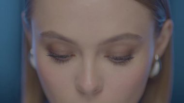 Yumuşak ışıklı, sakin ve anlamlı gözleri olan bir kadının aşırı yakın çekimi. İnce kamera hareketi ve pastel mavi arka plan yumuşak yüz ifadeleriyle rüya gibi, duygusal bir atmosfer yaratır..