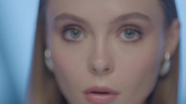 Güçlü ve odaklanmış bir ifadeye sahip, başını hafifçe eğmiş genç bir kadının yakın plan portresi. Yumuşak stüdyo ışıkları ve mavi arka plan altında yüksek çözünürlüklü görüntüler.