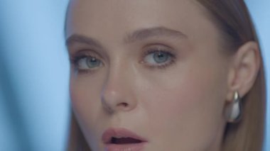 Güçlü ve odaklanmış bir ifadeye sahip, başını hafifçe eğmiş genç bir kadının yakın plan portresi. Yumuşak stüdyo ışıkları ve mavi arka plan altında yüksek çözünürlüklü görüntüler.