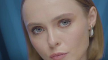 Güçlü ve odaklanmış bir ifadeye sahip, başını hafifçe eğmiş genç bir kadının yakın plan portresi. Yumuşak stüdyo ışıkları ve mavi arka plan altında yüksek çözünürlüklü görüntüler.